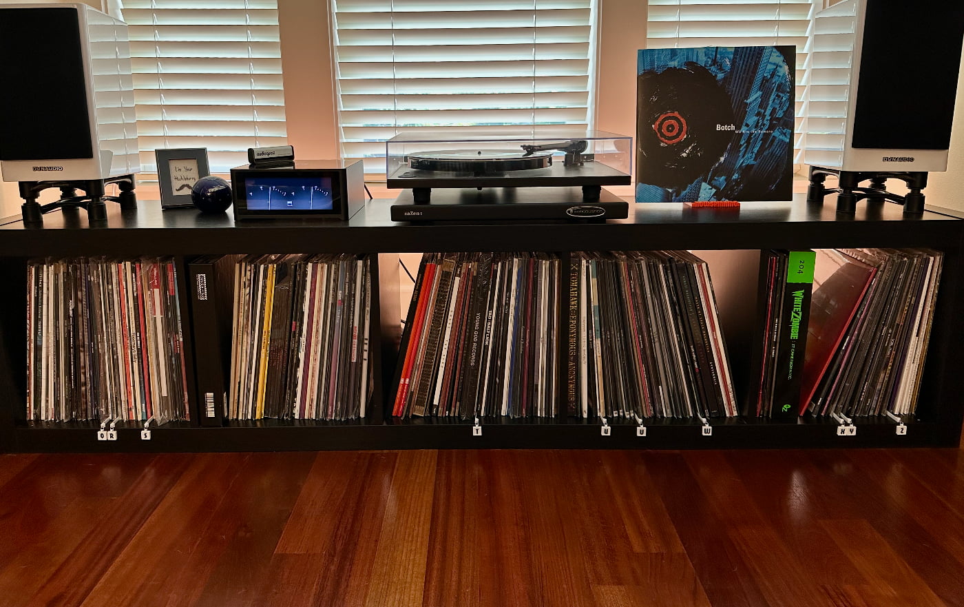 Small stereo setup including white speakers, an amplifier, and a turntable. Also shown are Super Heated products including letter shelf tags and the display stand. Underneath, five shelves filled with vinyl records.