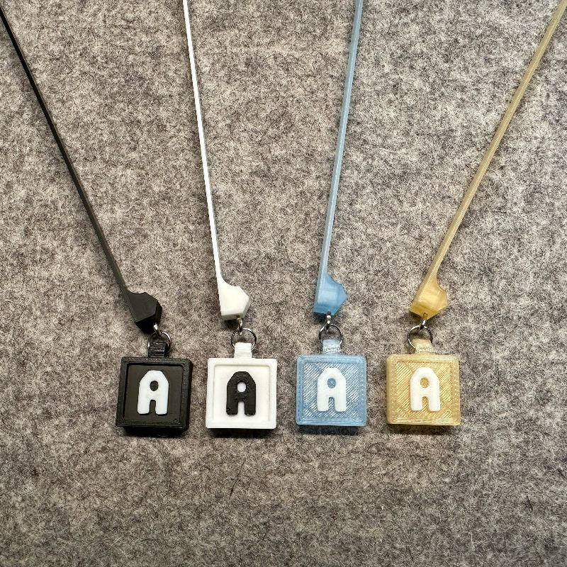 3D-printed letter tags shown in black, white, transparent blue, and transparent gold.